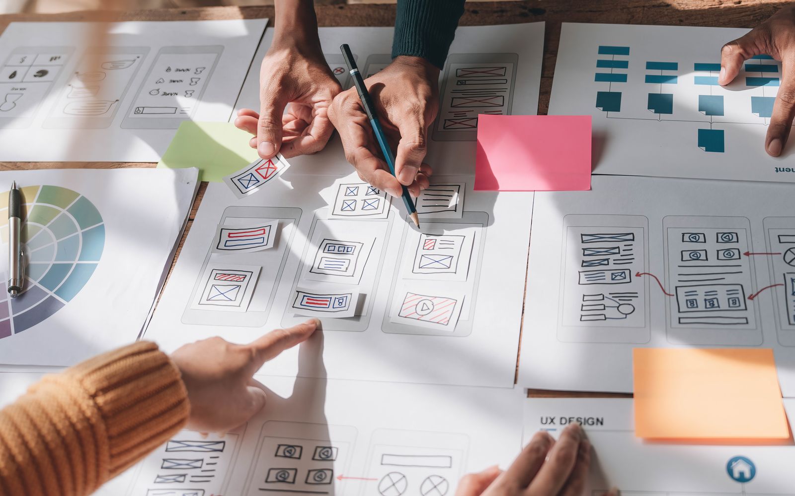 Manos de varias personas ordenan bocetos de pantallas de aplicación y diagramas de flujo sobre una mesa de trabajo