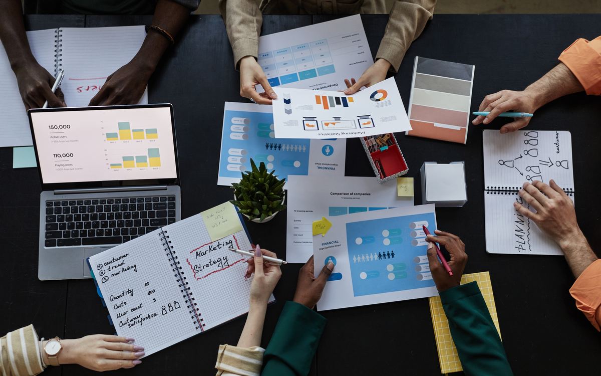 Vista superior de un equipo trabajando con laptop, cuadernos y gráficas impresas sobre una mesa oscura