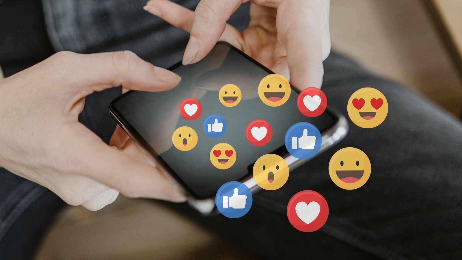 Manos sosteniendo un teléfono con emojis y reacciones flotando sobre la pantalla