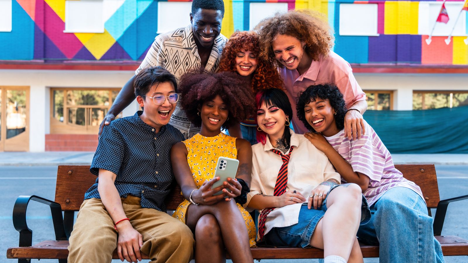 Grupo de amigos sonriendo mientras miran un teléfono sentados en una banca frente a un mural colorido
