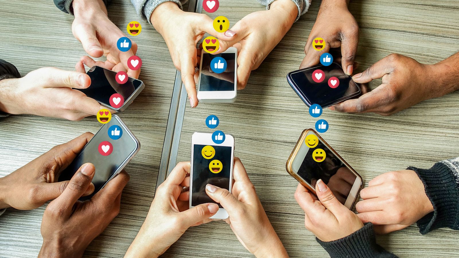 Manos alrededor de una mesa usando teléfonos con íconos de reacciones flotando
